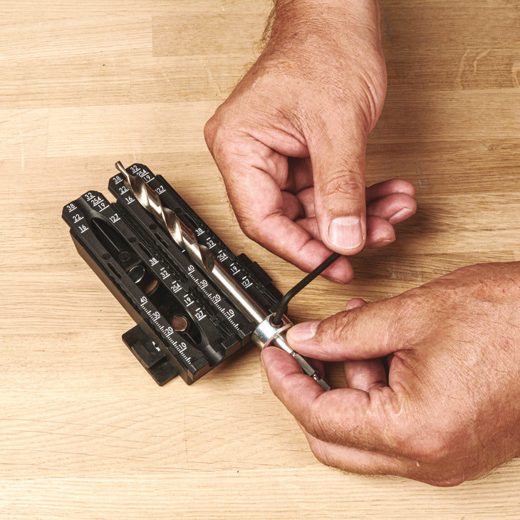 Close-up of a person's hands using a black and silver drill bit size guide on a light wood surface.